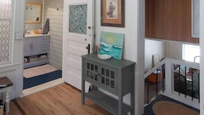 A room with a wooden floor, a grey bench, a door with a 'Welcome' sign, and a small table with a bowl. - Lock`s Nest on Peaks Island Maine (Portland)