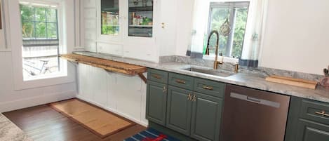 A kitchen with wooden cabinets, a built-in oven, a stainless steel dishwasher, and a window with a view of greenery.