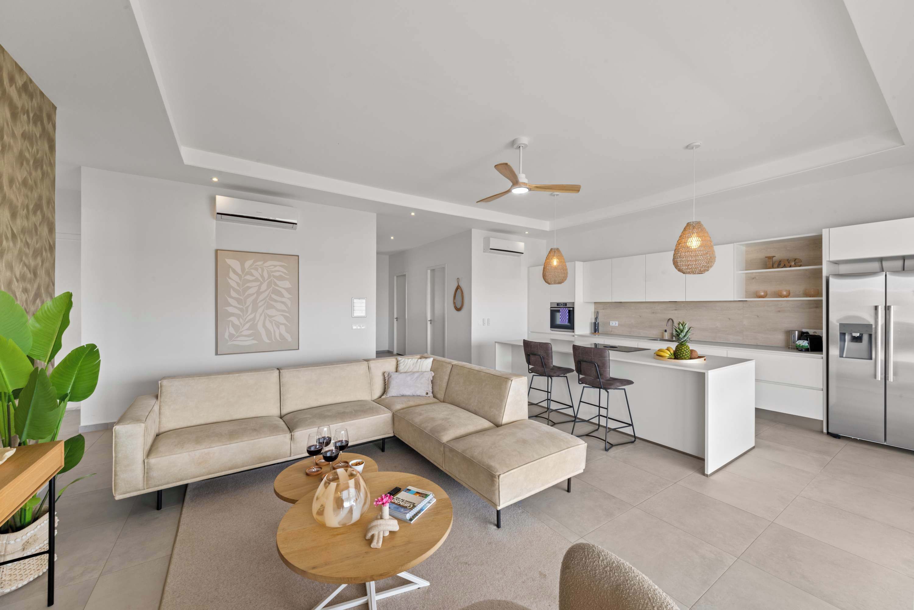 A modern living room with a sofa, coffee table, and a kitchen area with a refrigerator, microwave, and bar stools.