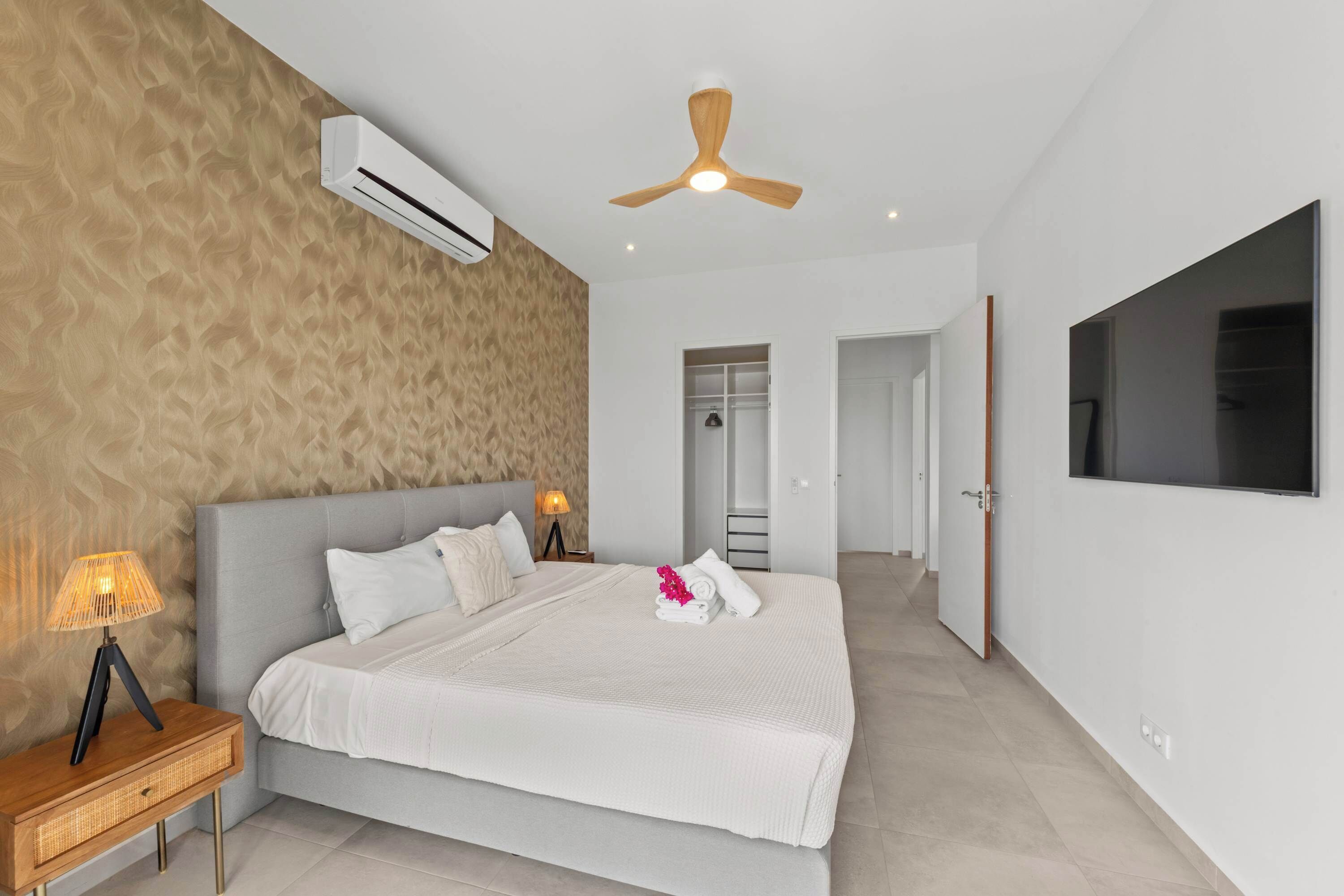 A modern bedroom with a bed, bedside lamps, a ceiling fan, and a TV mounted on the wall.