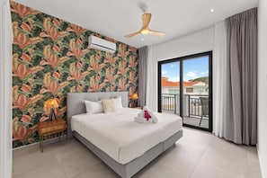 Ein Hotelzimmer mit einem Bett, einem Deckenventilator, einem Fenster mit Blick auf Gebäude und einer Wand mit einem tropischen Blattmuster.