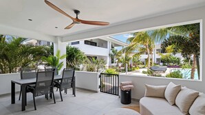 Un patio moderno al aire libre con mesa, sillas, ventilador de techo y vistas a una piscina y follaje.