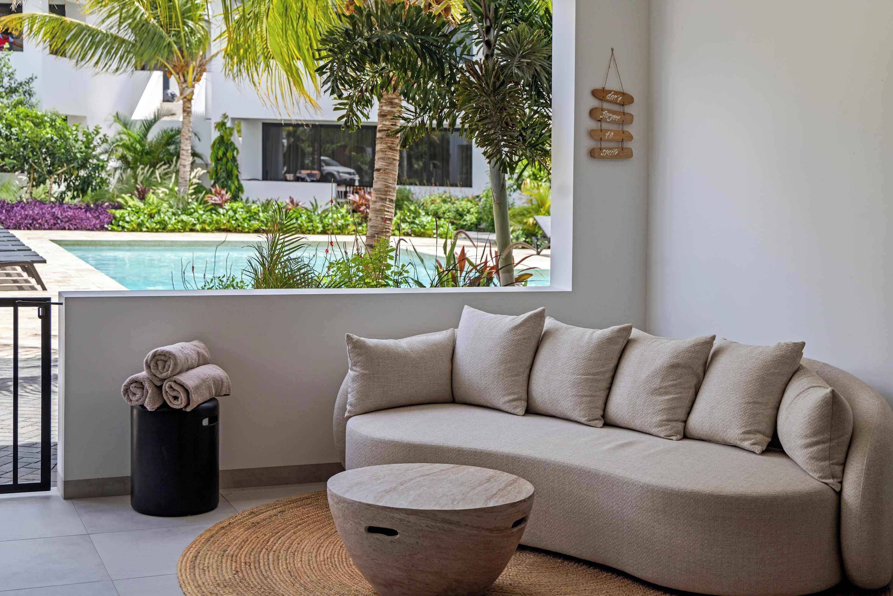 A modern living room with a beige sofa, a woven coffee table, and a view of a pool and greenery.