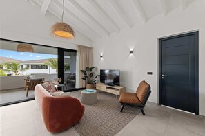 A modern living room with a flat-screen TV, comfortable seating, and a large sliding glass door leading to a balcony. - Sunset Pearl Curaçao | Pool & Free Parking (Mahaai)