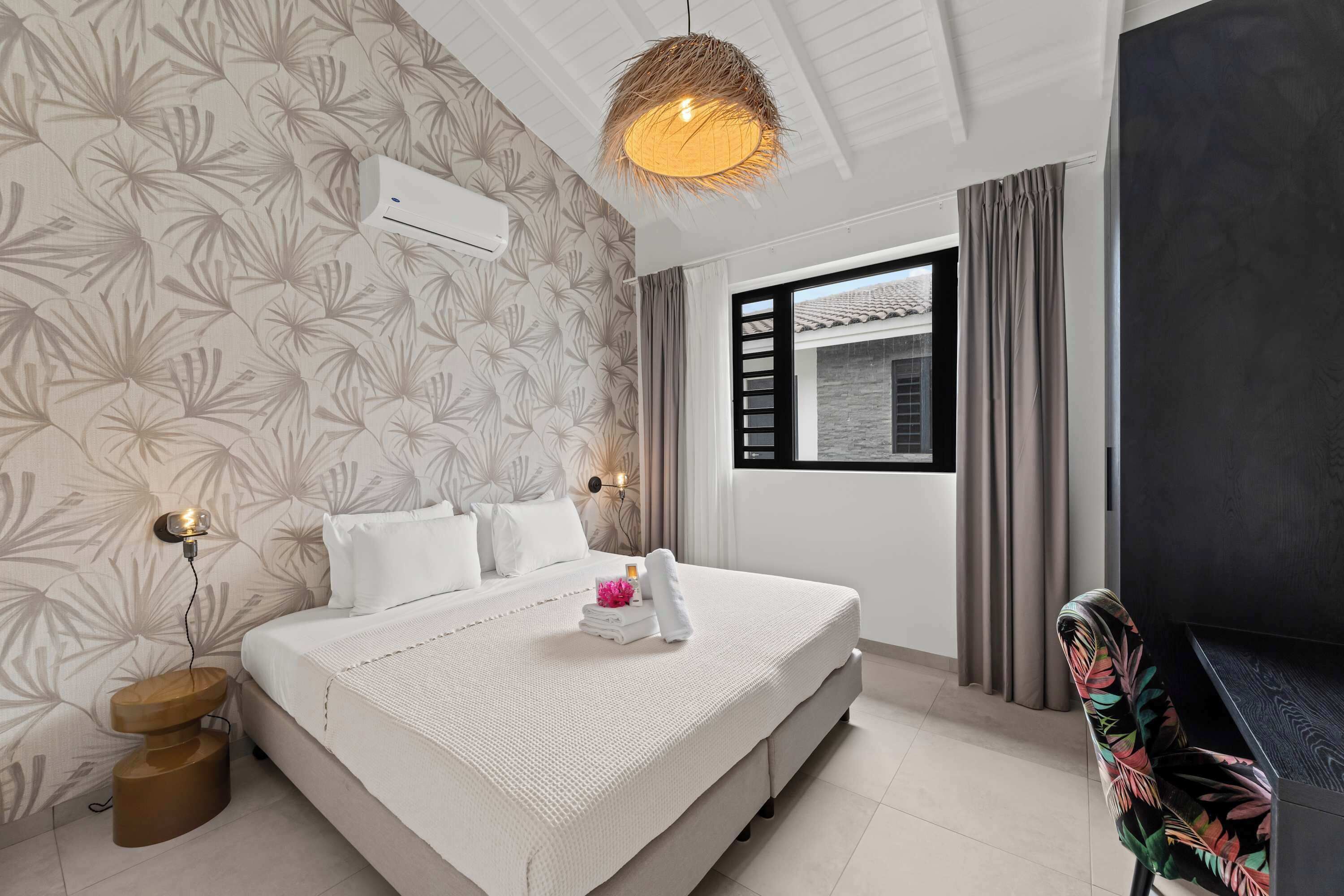 A hotel room with a bed, a bedside lamp, a wall-mounted air conditioner, a window with curtains, and a view of the outside.