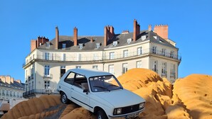 Ein weißes Auto steht in einer sandigen Umgebung, im Hintergrund ist ein Gebäude zu sehen.
