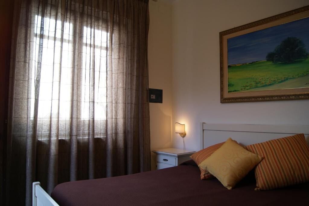 A bedroom with a bed, a nightstand, a lamp, a painting on the wall, and a window with curtains.