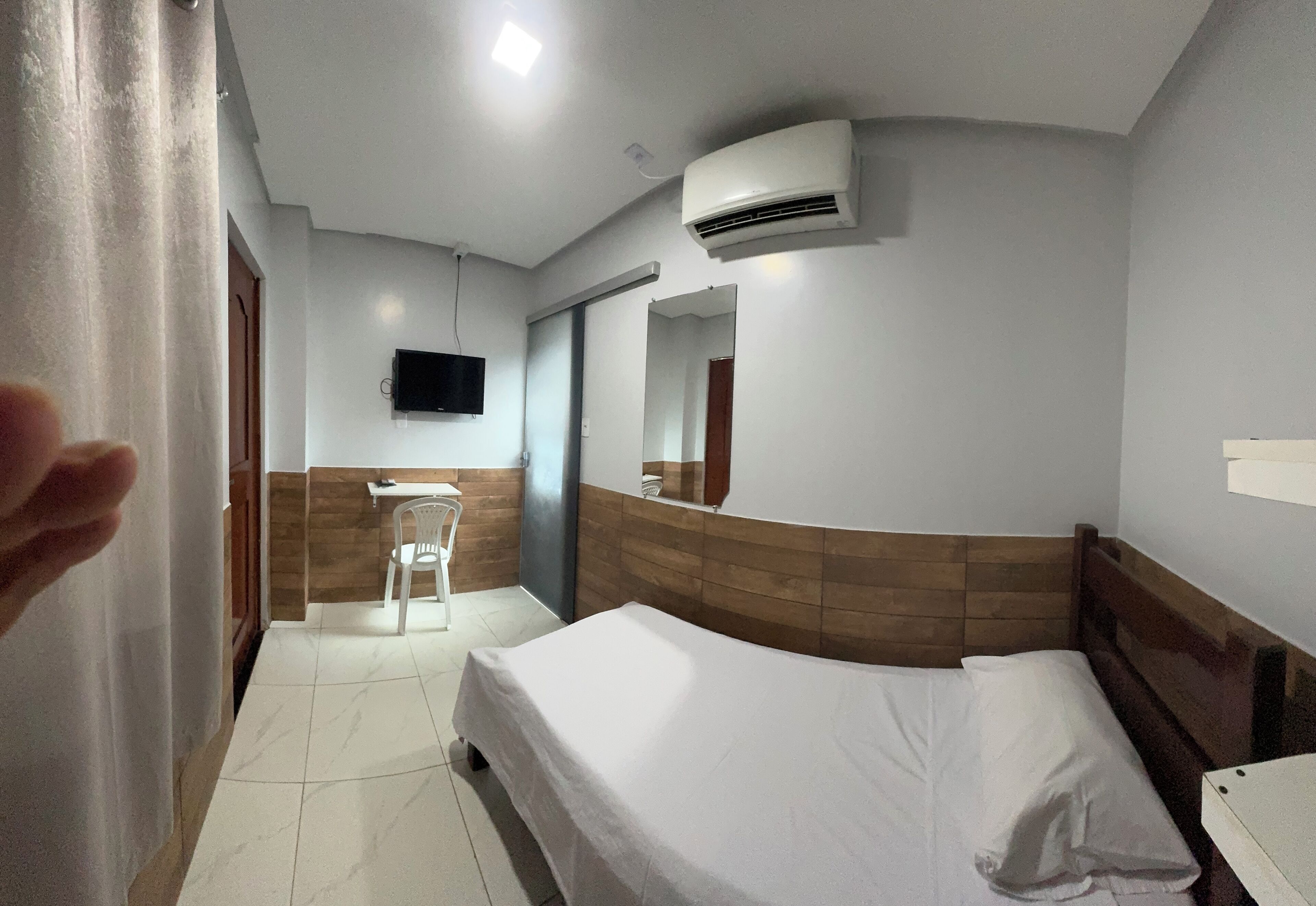 Ein Hotelzimmer mit einem Bett, einem kleinen Tisch, einem Sessel, einem an der Wand befestigten Fernseher und einer Klimaanlage.