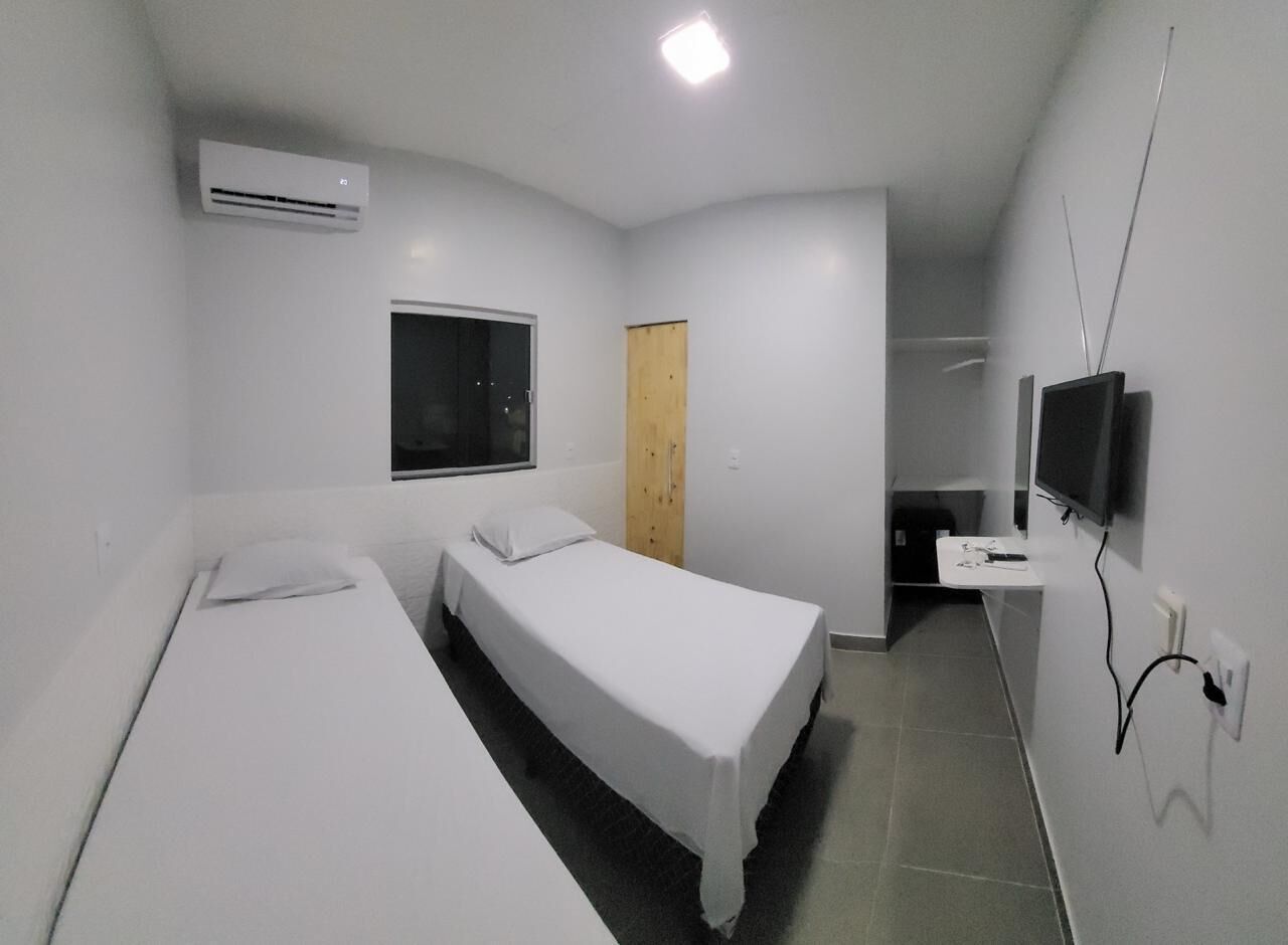 A small, clean room with two single beds, a window, a wooden door, a TV mounted on the wall, and an air conditioning unit.