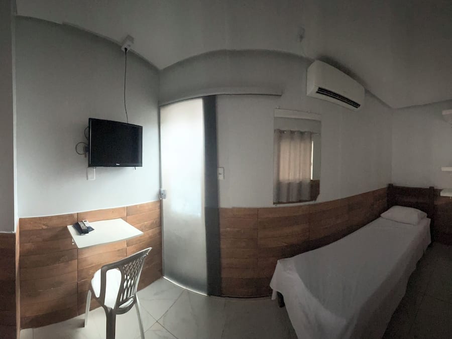 Una camera compatta con un letto, un piccolo tavolo, una sedia, una TV a parete e una finestra con tende.