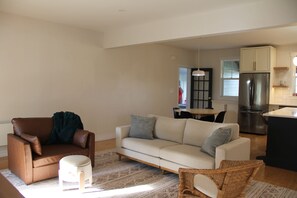 A living room with a brown leather chair, a white sofa, and a small stool. There is a dining table with chairs in the background.