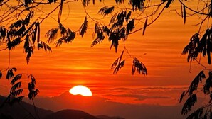 A sunset with the sun partially obscured by clouds, silhouetted trees, and a mountain range in the background.