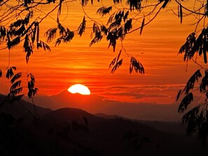 A sunset with the sun partially obscured by clouds, silhouetted trees, and a mountain range in the background. - El Mirador Gualán (Gualán)