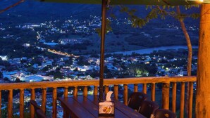 An outdoor dining area with a table and chairs under a green umbrella, overlooking a cityscape at dusk.