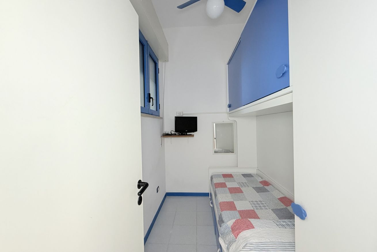 A small, narrow room with a single bed, a television mounted on the wall, and a ceiling fan.
