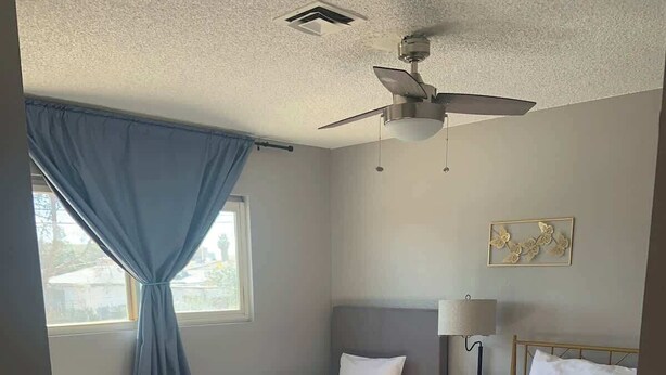 Una camera da letto con due letti, una ventola a soffitto, una finestra con tende, una lampada da comodino e un piccolo tavolo.