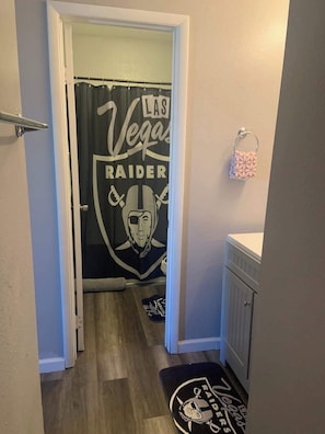 Ein Flur mit einer Duschvorhangs, der das Logo der Vegas Raiders zeigt, einem kleinen Handtuchhalter mit einem gemusterten Handtuch und einer Fußmatte mit demselben Logo.