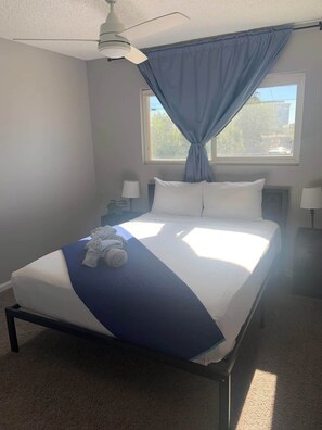 Ein Einzelbett mit blauer und weißer Bettdecke, ein Deckenventilator, ein Fenster mit blauen Vorhängen und ein Nachttisch mit einer Lampe.