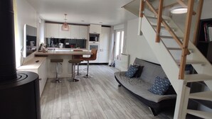 Una cocina moderna con una isla central, un área de comedor con taburetes de bar, un sofá con cojines estampados y una escalera con barandilla de madera.