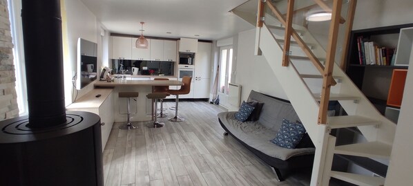 A modern kitchen with a central island, a dining area with bar stools, a sofa with patterned cushions, and a staircase with a wooden railing.