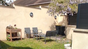 Un patio con mesa y sillas, un pequeño jardín y un edificio con tejado de baldosas.