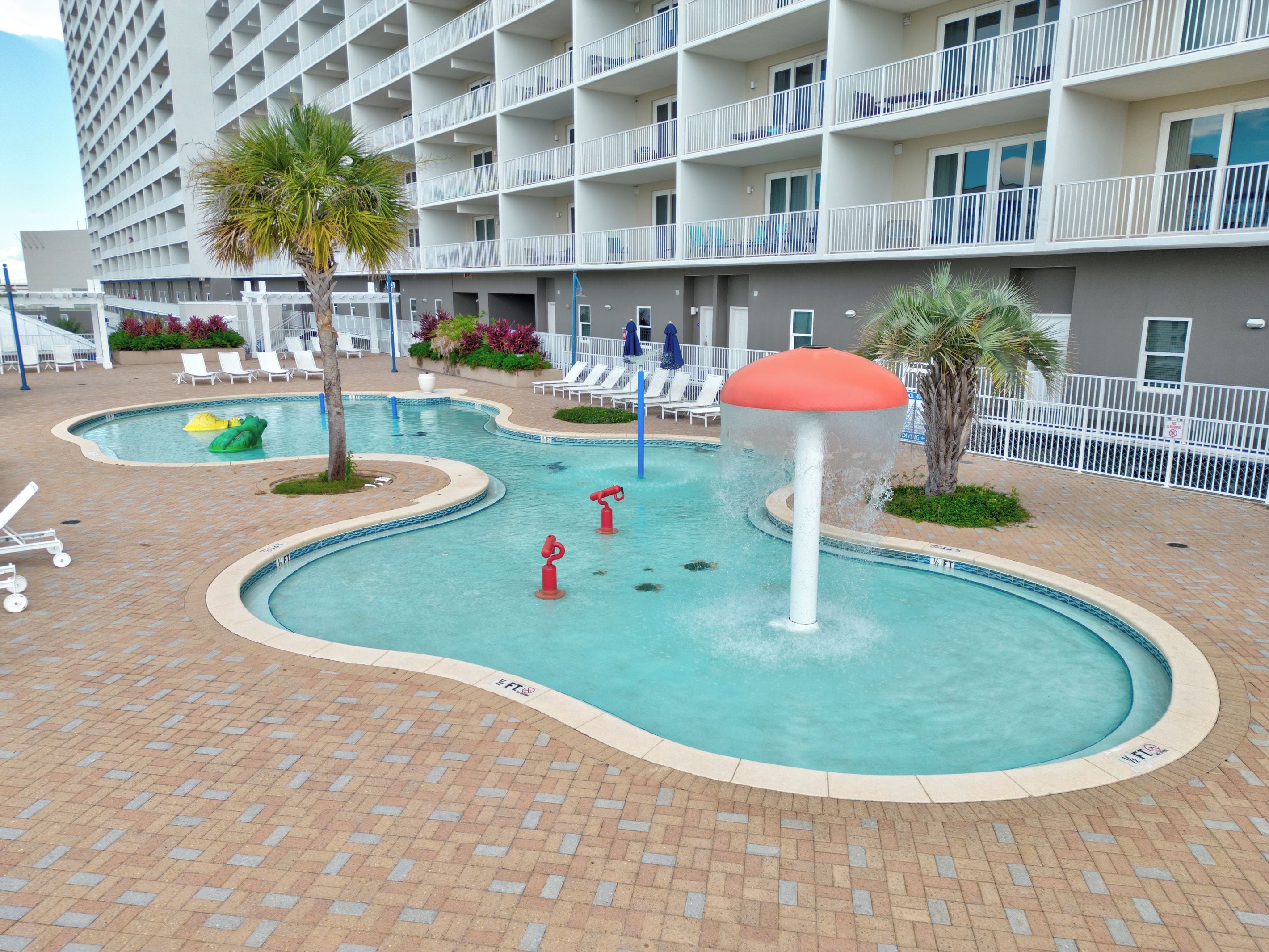 A swimming pool area with multiple pools, lounge chairs, and a building with balconies in the background.