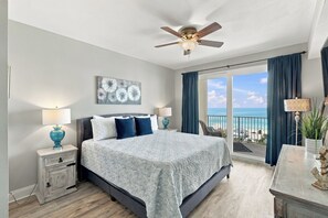 A bedroom with a bed, bedside tables, a ceiling fan, a view of the ocean, and a large window with curtains. - Ocean View Condo | 5 Pools & Beach (Panama City Beach)