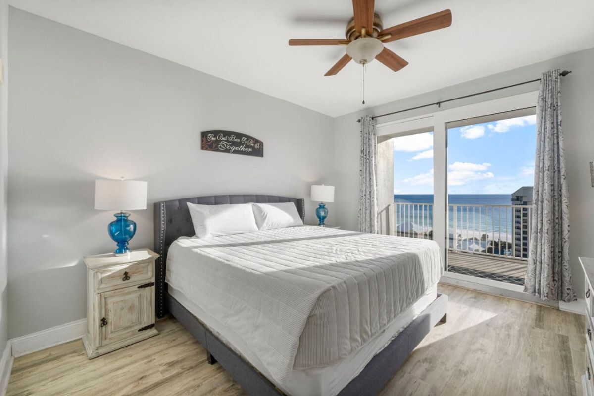 Un dormitorio con cama, mesitas de noche, ventilador de techo, ventana con cortinas y balcón con vistas.