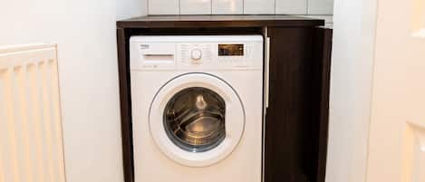 Una lavadora blanca de carga frontal, compacta, ubicada en un rincón con un mueble de madera oscura encima.