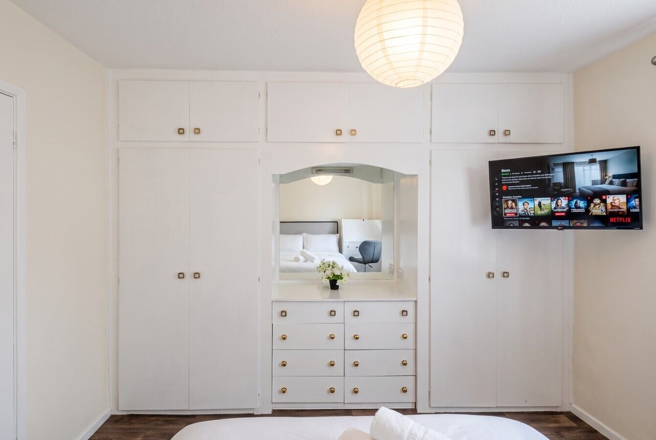 A bedroom with a bed, a dresser with drawers, a mirror, a television mounted on the wall, and a ceiling light fixture.