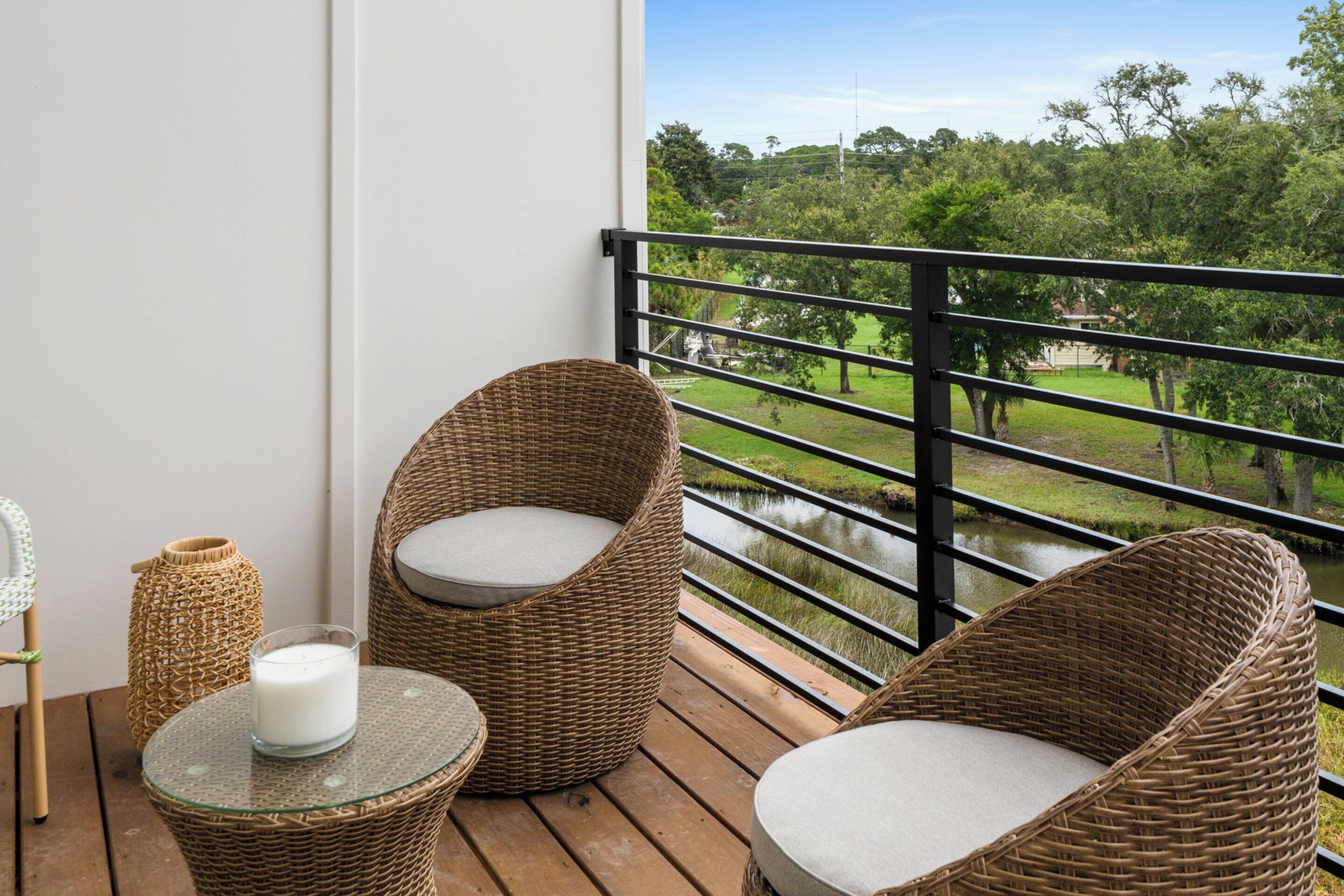 A balcony with wicker chairs and a glass table, overlooking a grassy area with a water body.
