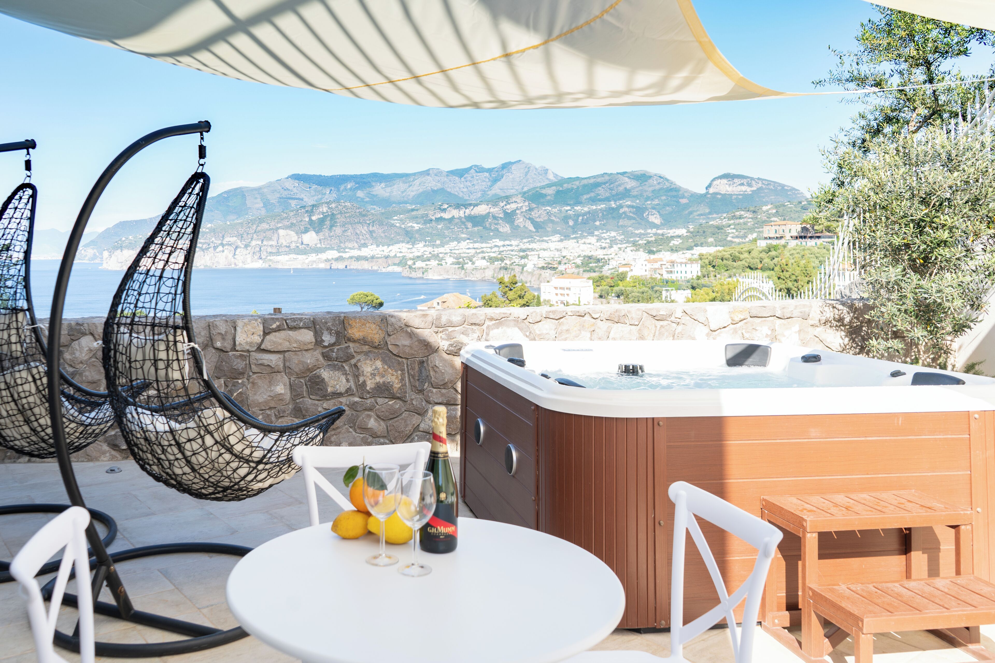 Un patio con hidromasaje, dos sillas colgantes, una mesa con vasos y limones, y una vista panorámica de un pueblo costero y montañas.
