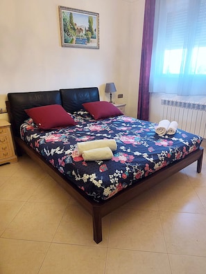 Ein Bett mit floralem Überwurf, zwei rote Kissen und zwei zusammengerollte Handtücher.
