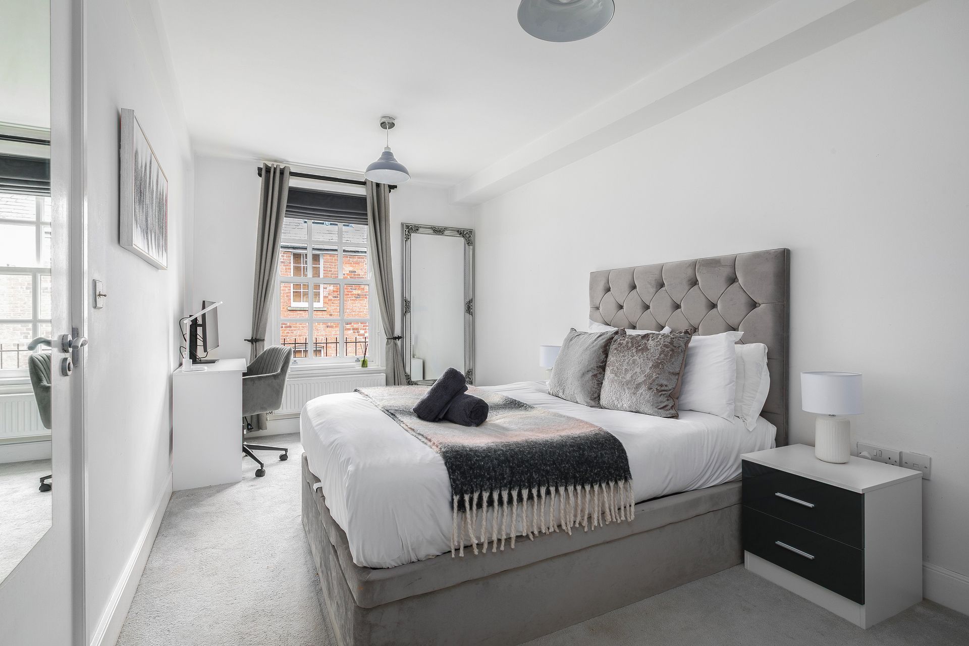 Ein modernes Schlafzimmer mit einem großen Bett, Nachttischen, einem Sessel und Blick auf ein Backsteingebäude durch ein Fenster.