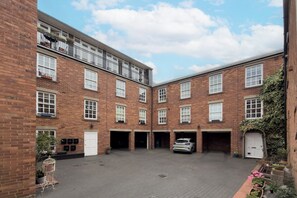 A courtyard with a car parked in a garage, surrounded by brick buildings with multiple windows and balconies. - Stylish 2BDR 2BA Flat with Chic Interiors (West Midlands)