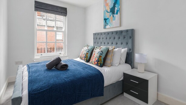 A bedroom with a bed, a bedside table, a window, and a painting on the wall. - Stylish 2BDR 2BA Flat with Chic Interiors (West Midlands)