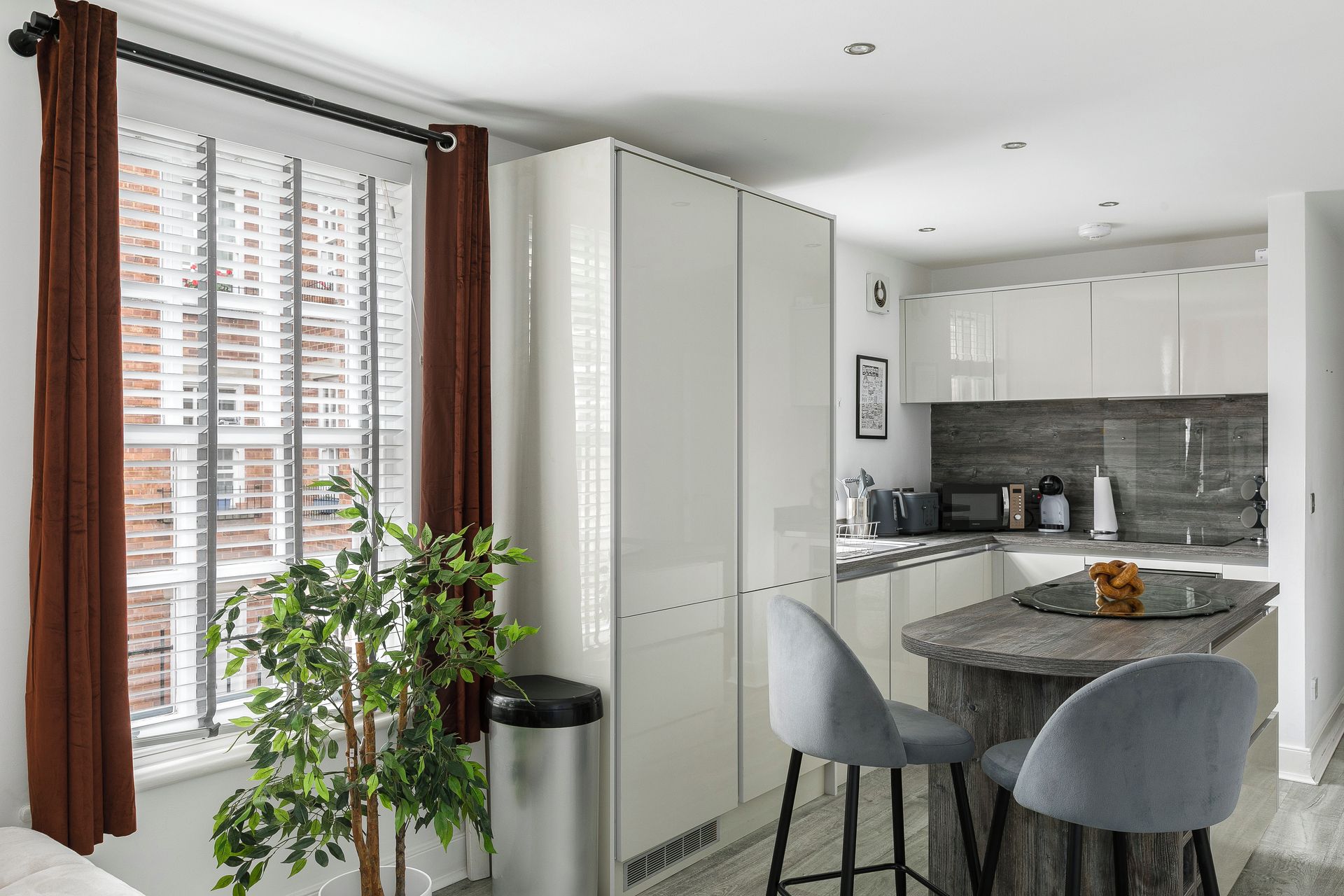 Una cocina moderna con armarios blancos, una zona de comedor con dos sillas grises y vistas a un paisaje urbano a través del ventanal.