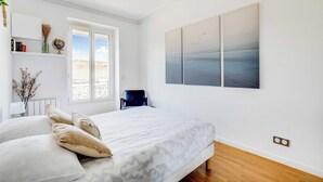 Minimalistinen makuuhuone, jossa on sänky, tuoli, ikkuna ja taideteos seinällä.