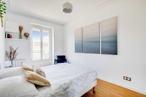 Ein schlichter Schlafzimmerbereich mit Bett, Sessel, Fenster und einem Kunstwerk an der Wand.