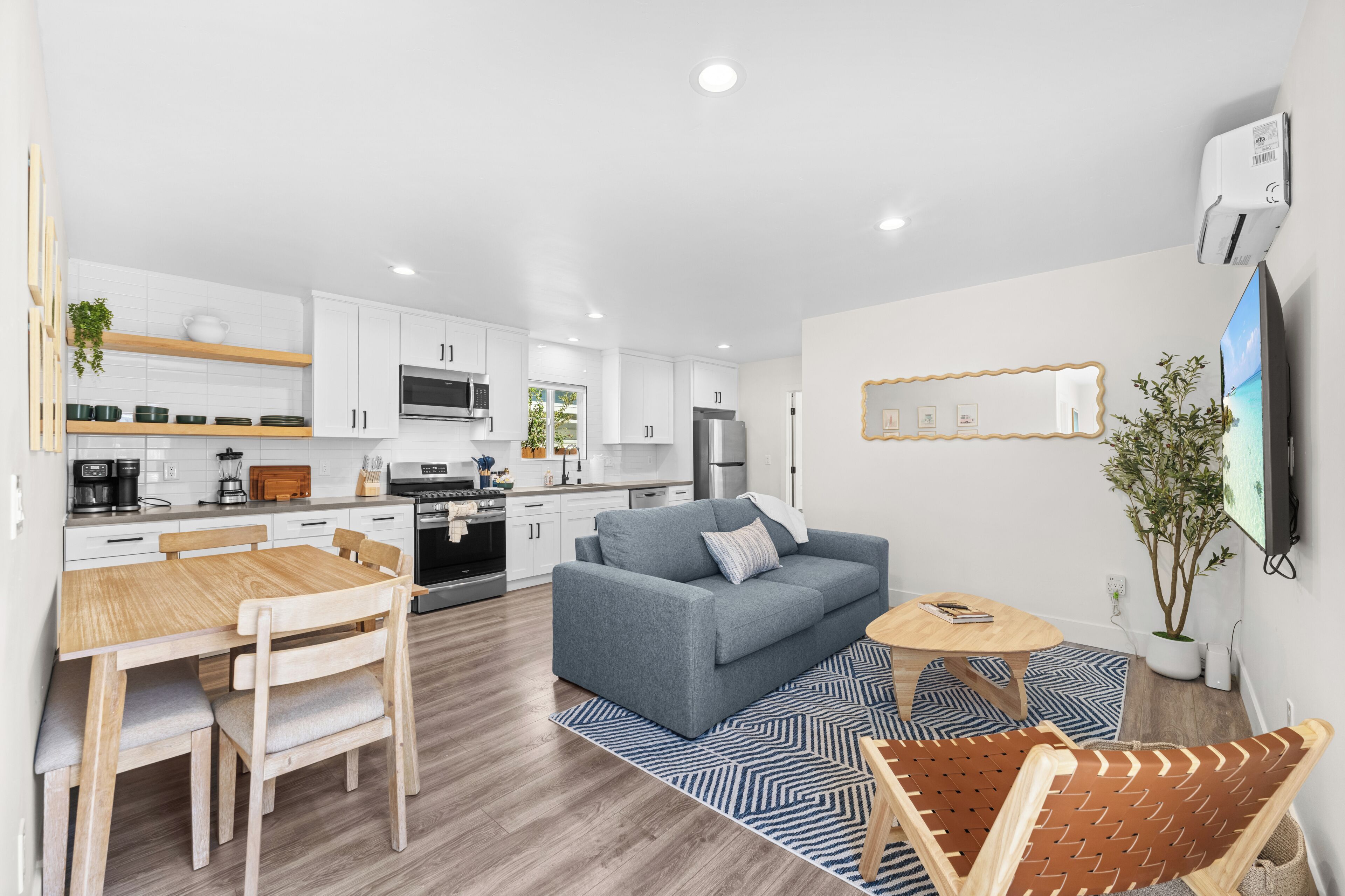 A modern kitchen with white cabinets, a dining area with a wooden table and chairs, a grey sofa, and a TV mounted on the wall.