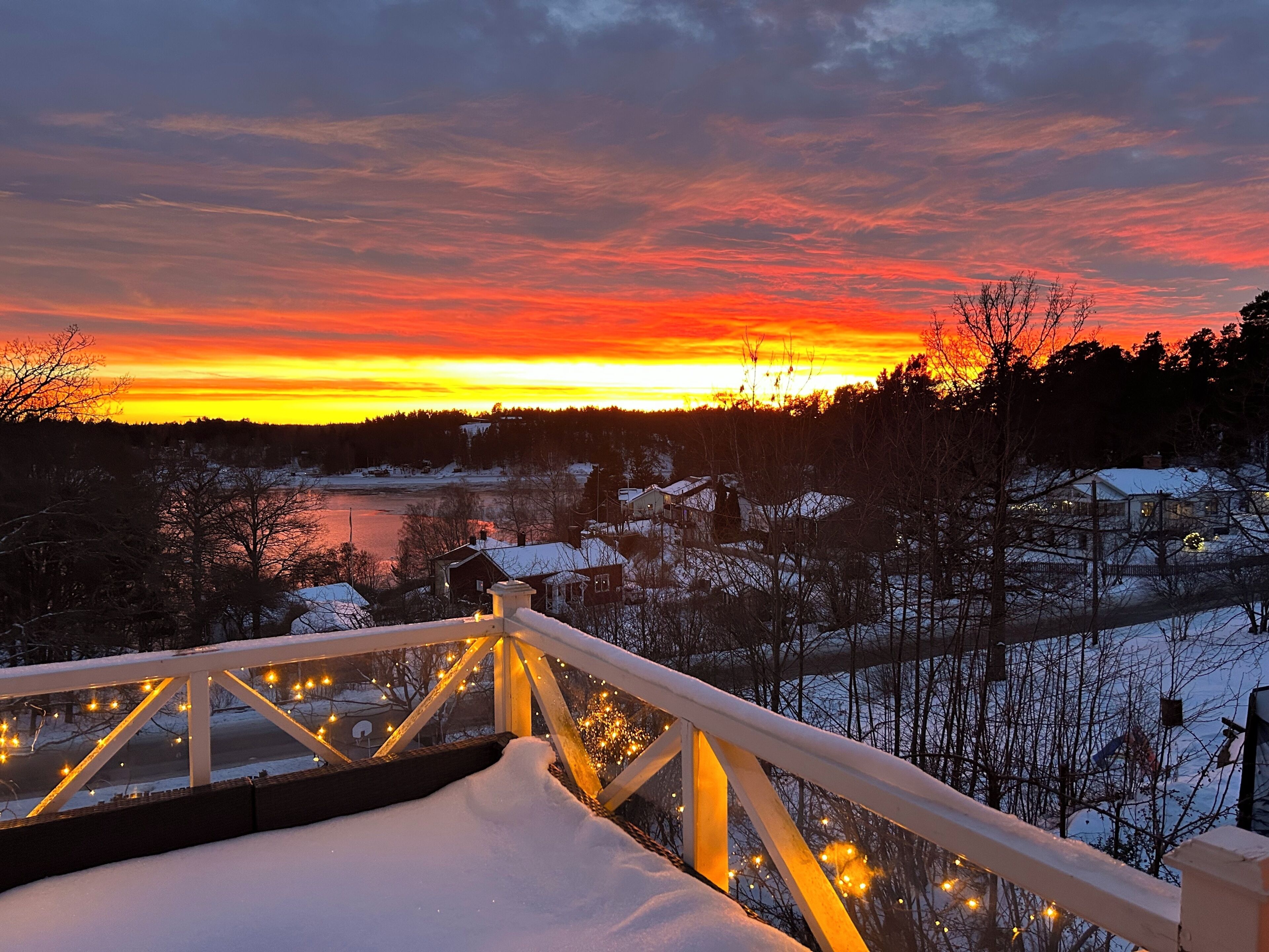 Ein mit Schnee bedeckter Balkon mit Geländer und Beleuchtung, der auf eine Winterlandschaft mit Sonnenuntergang blickt.