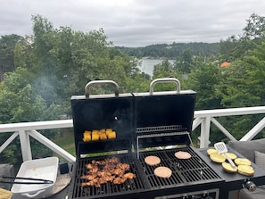 A barbecue grill with corn on the cob and hamburgers, set on a balcony with a view of a lake and forest.