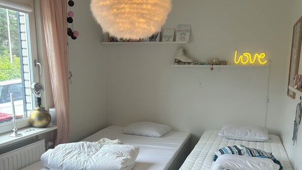 Ein Schlafzimmer mit zwei Betten, einer runden Leuchte, einem Fenster mit Vorh ängen und einem Neon-Schild an der Wand.
