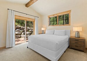 A bedroom with a large bed, a nightstand, a window with a view of greenery, and a sliding door leading to an outdoor area. - Perfect Family Getaway: 4BR with Stunning Alpine Views (Keystone)
