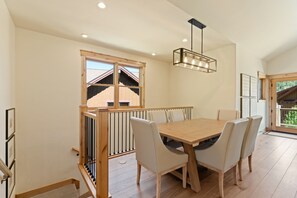 A dining area with a wooden table and chairs, a hanging light fixture, and a view of a building through a window. - Perfect Family Getaway: 4BR with Stunning Alpine Views (Keystone)