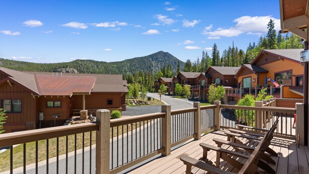 A wooden deck with outdoor seating, overlooking a mountain and a road with multiple cabins. - Perfect Family Getaway: 4BR with Stunning Alpine Views (Keystone)