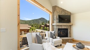 A modern living room with a stone fireplace, comfortable seating, and a large window overlooking a mountainous landscape. - Perfect Family Getaway: 4BR with Stunning Alpine Views (Keystone)
