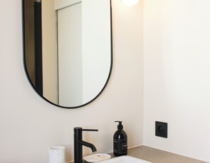 A bathroom with a round mirror, a black faucet, a soap dispenser, and a white cup. - Charming 1-bedroom flat in the 9th (Paris)