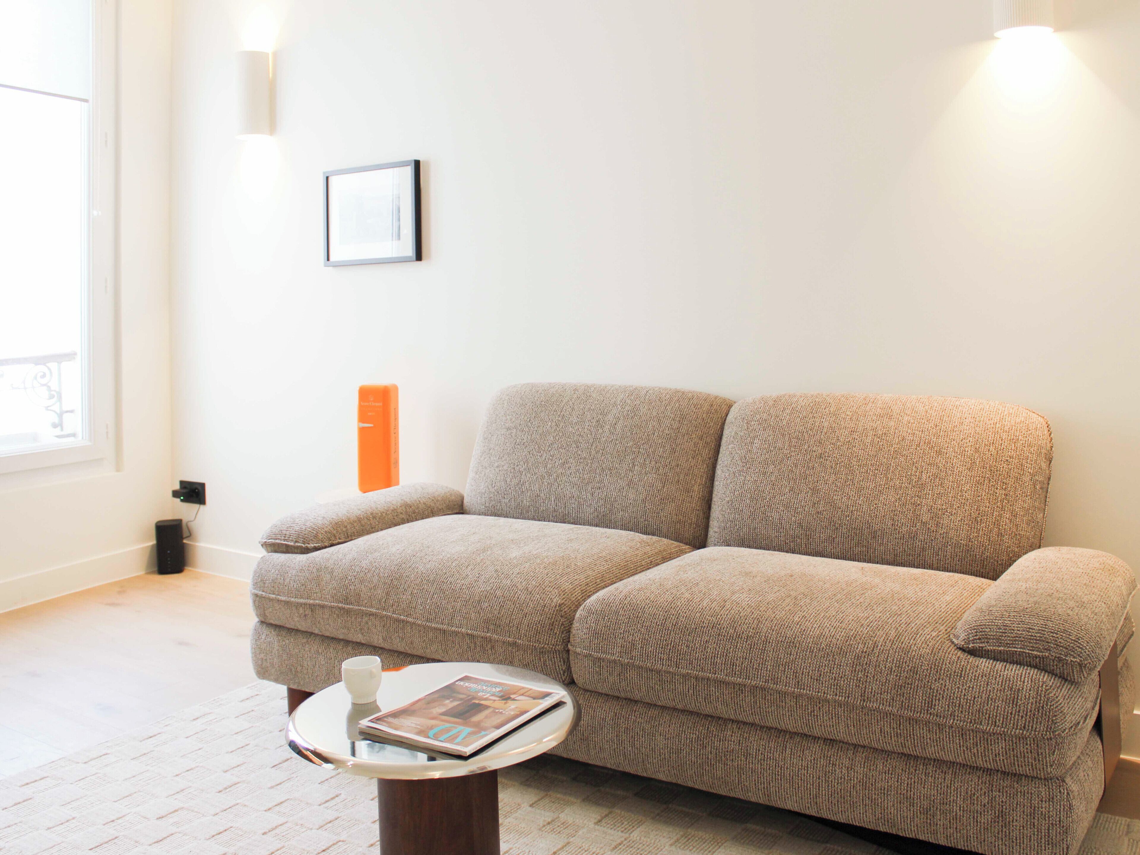 Een woonkamer met een beige bank, een klein rond salontafeltje en een ingelijst schilderij aan de muur.