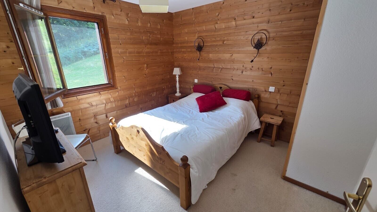 Une chambre à coucher avec un lit en bois, un téléviseur posé sur un meuble, une petite table de chevet et une fenêtre donnant sur un paysage verdoyant.